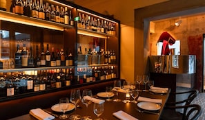 Zona de bar con estantes llenos de botellas de vino, copas y mesas de comedor ordenadas.