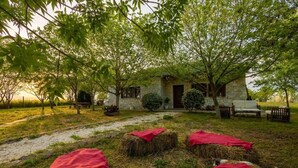 Un rustico edificio in pietra con un vialetto in pietra che vi conduce, circondato da alberi e balle di fieno con drappi rossi.