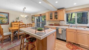 A kitchen with wooden cabinets, a central island with a stove, a dining area with chairs, and a window with a view of trees. - Two Story Townhome with Hot Tub, Walk to Lake (Tahoe City)