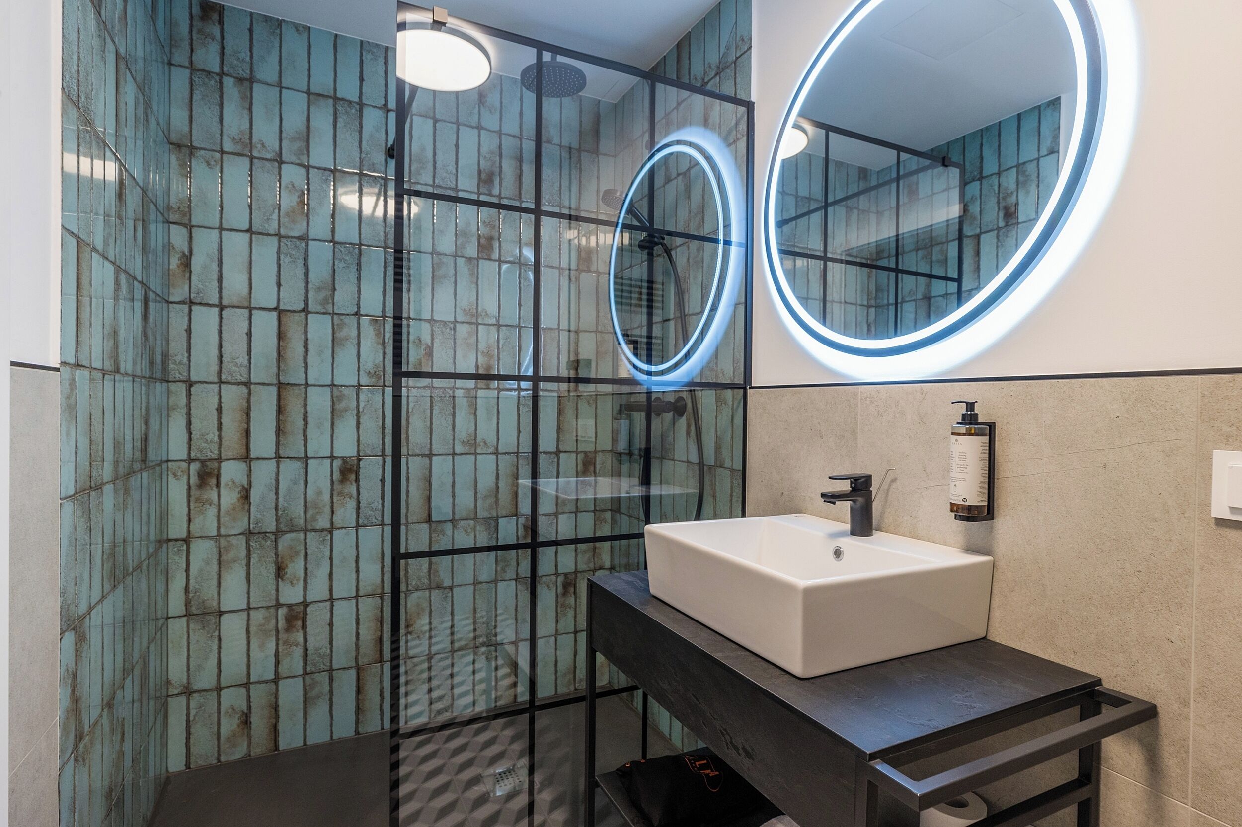 A modern bathroom with a glass shower enclosure, a white sink, and a round mirror with a blue light.