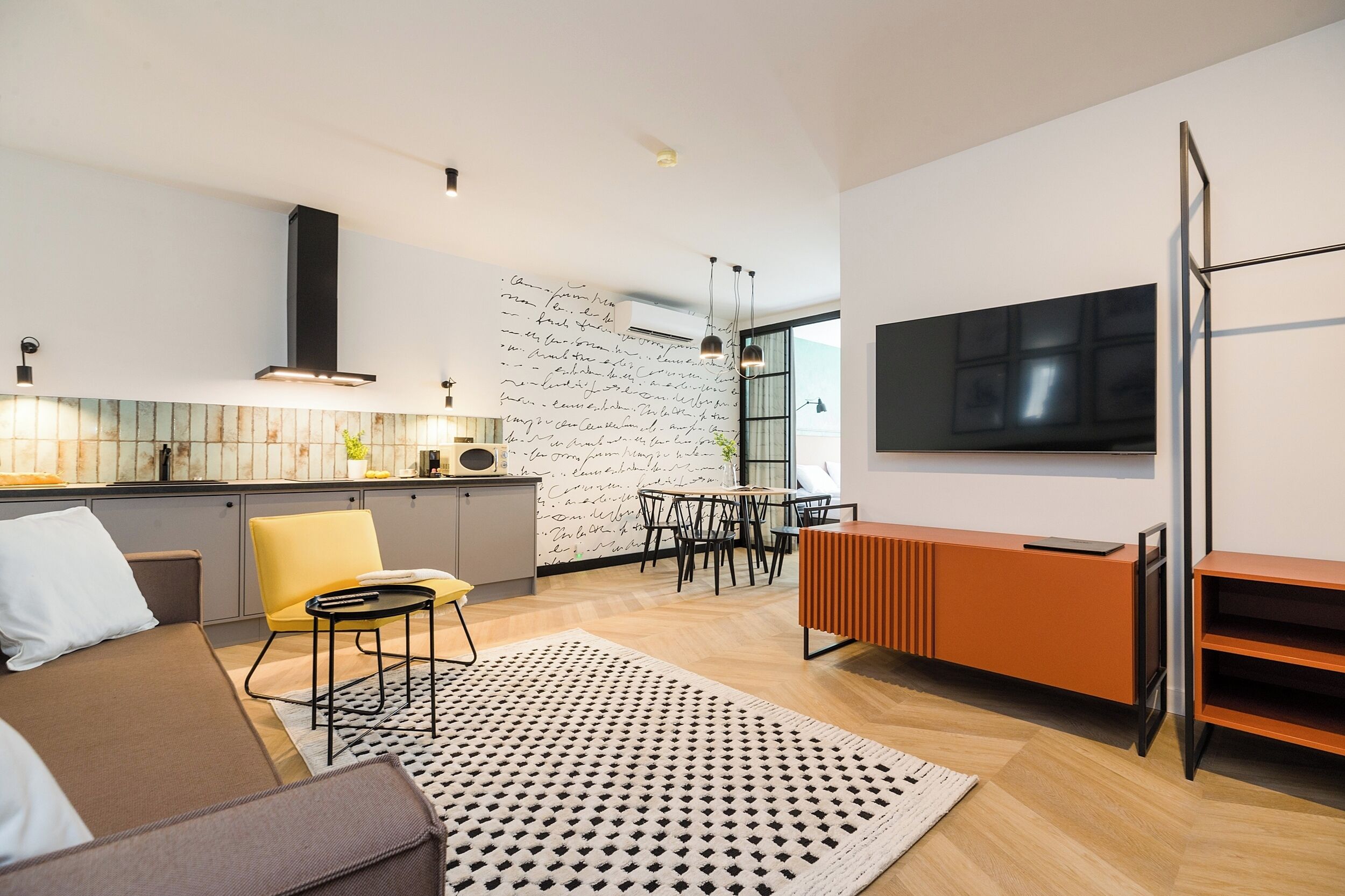 Un salon moderne comprenant un canapé, un coin repas, une cuisine dotée d’une grande fenêtre et un téléviseur fixé au mur.