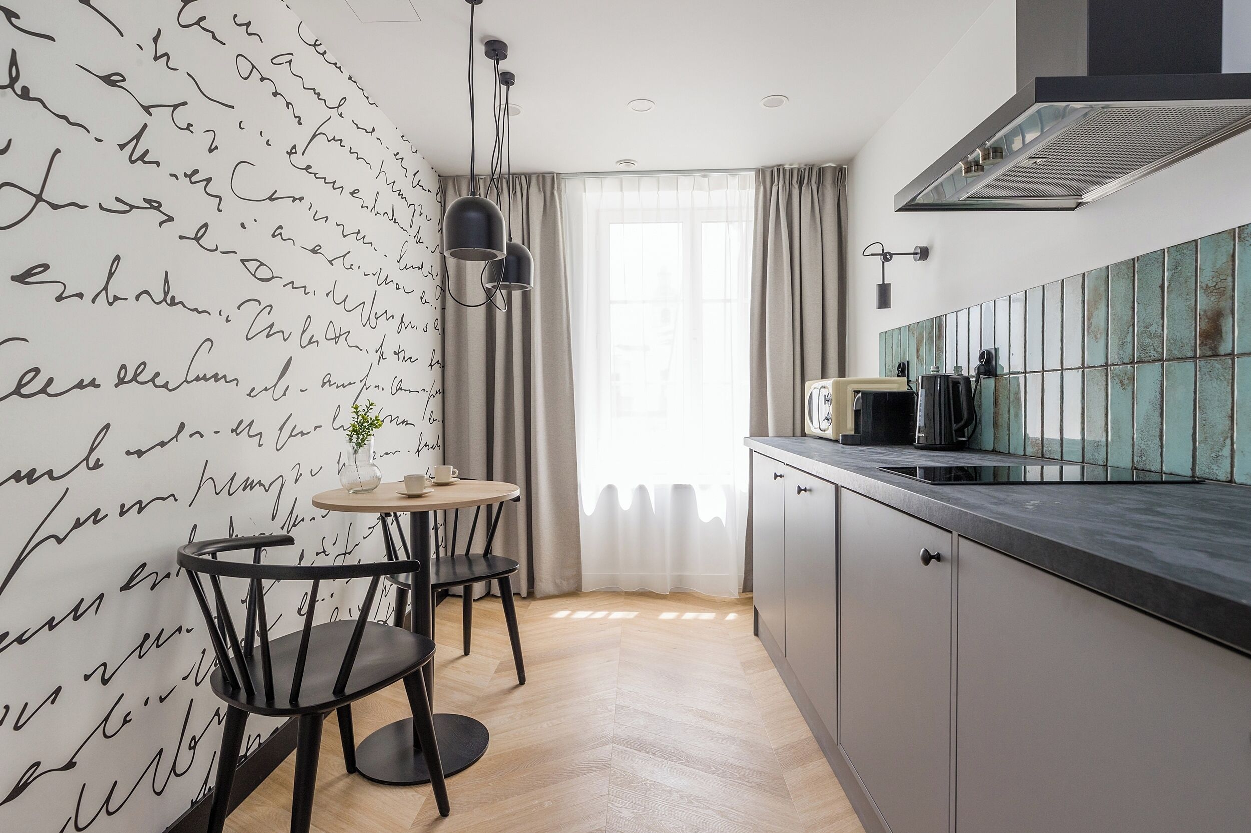 A modern kitchen with a herringbone wood floor, a round dining table with two chairs, a pendant light, and a view of the outdoors through a large window with sheer curtains.