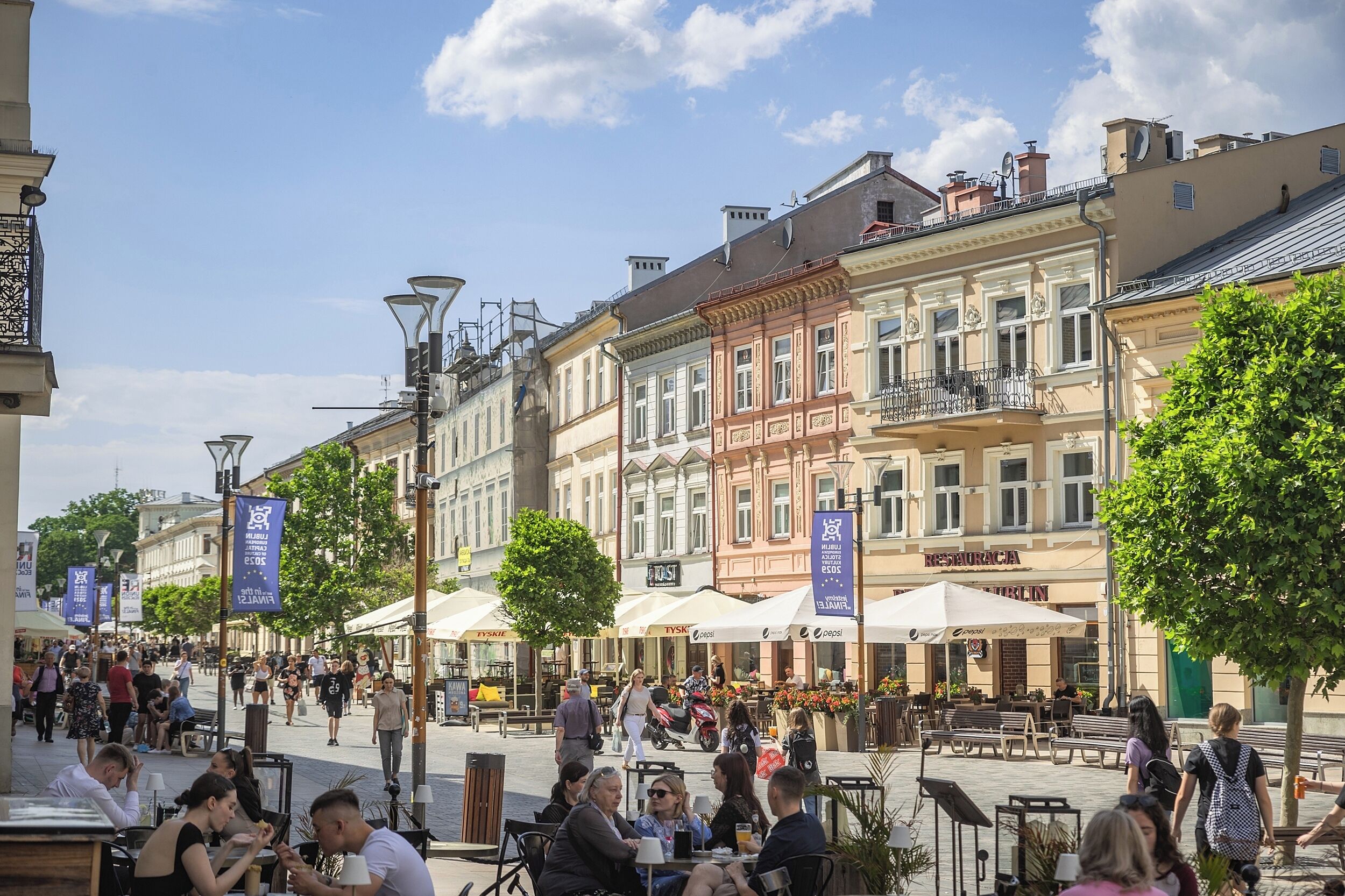 A bustling outdoor café area with tables and chairs, surrounded by historic buildings with balconies and large windows.