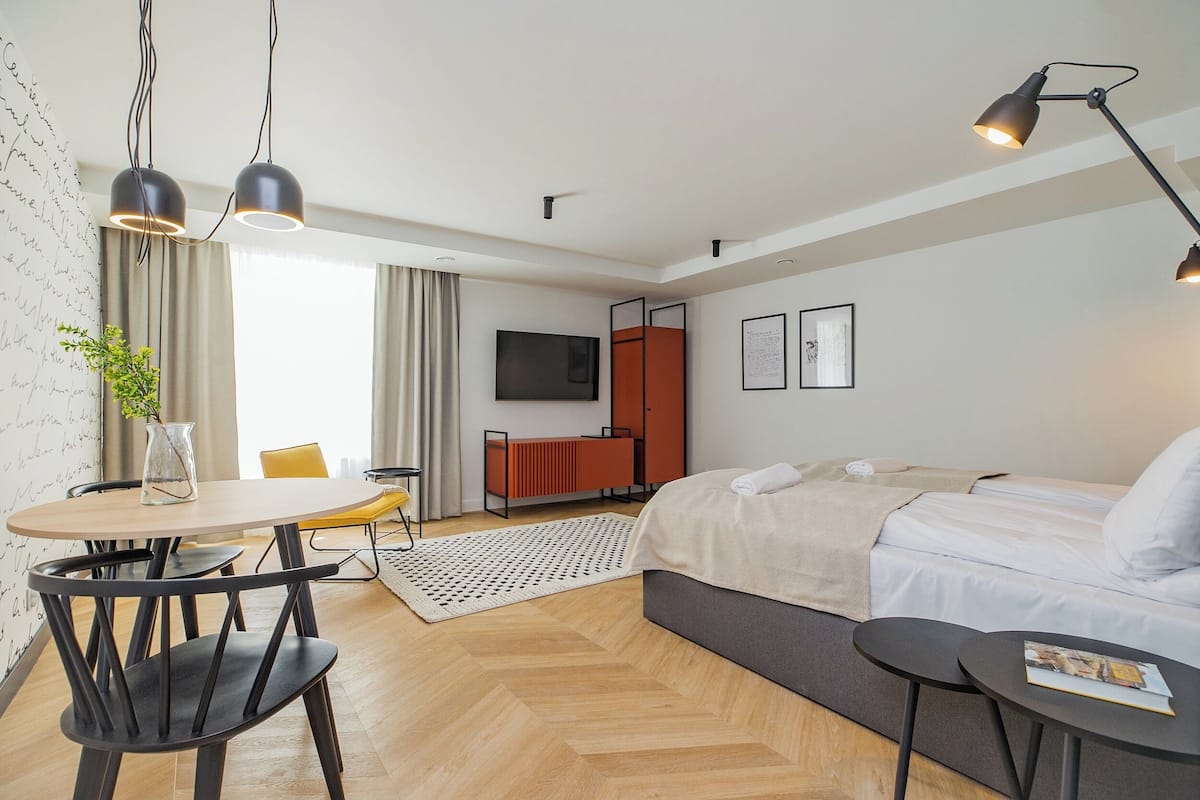 A modern hotel room with a bed, bedside table, and a TV. There is a dining area with a table and chairs, and a small plant on the table.
