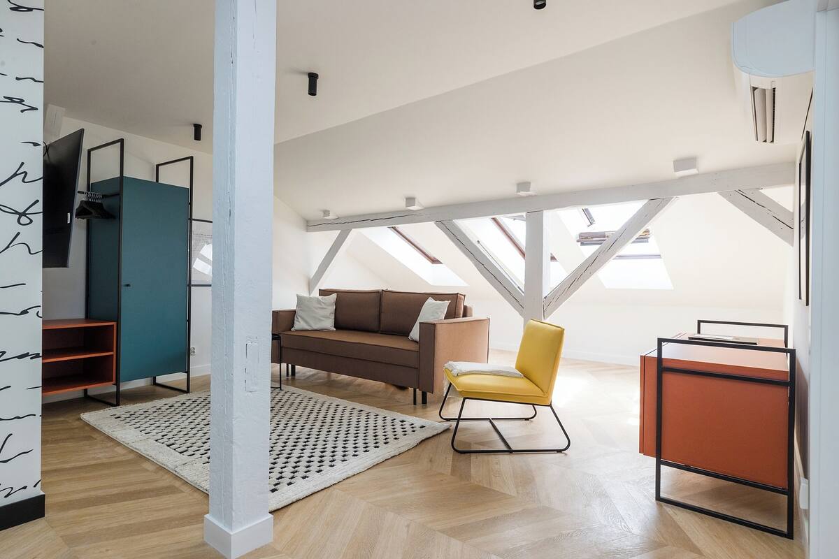 A modern loft bedroom with a high ceiling, a brown sofa, a yellow chair, and a small table.