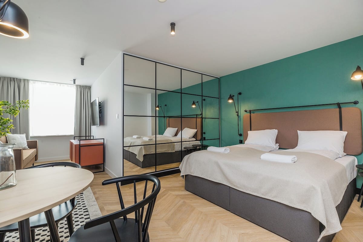 A modern hotel room with a large bed, a desk with a chair, a small table, and a large mirror.