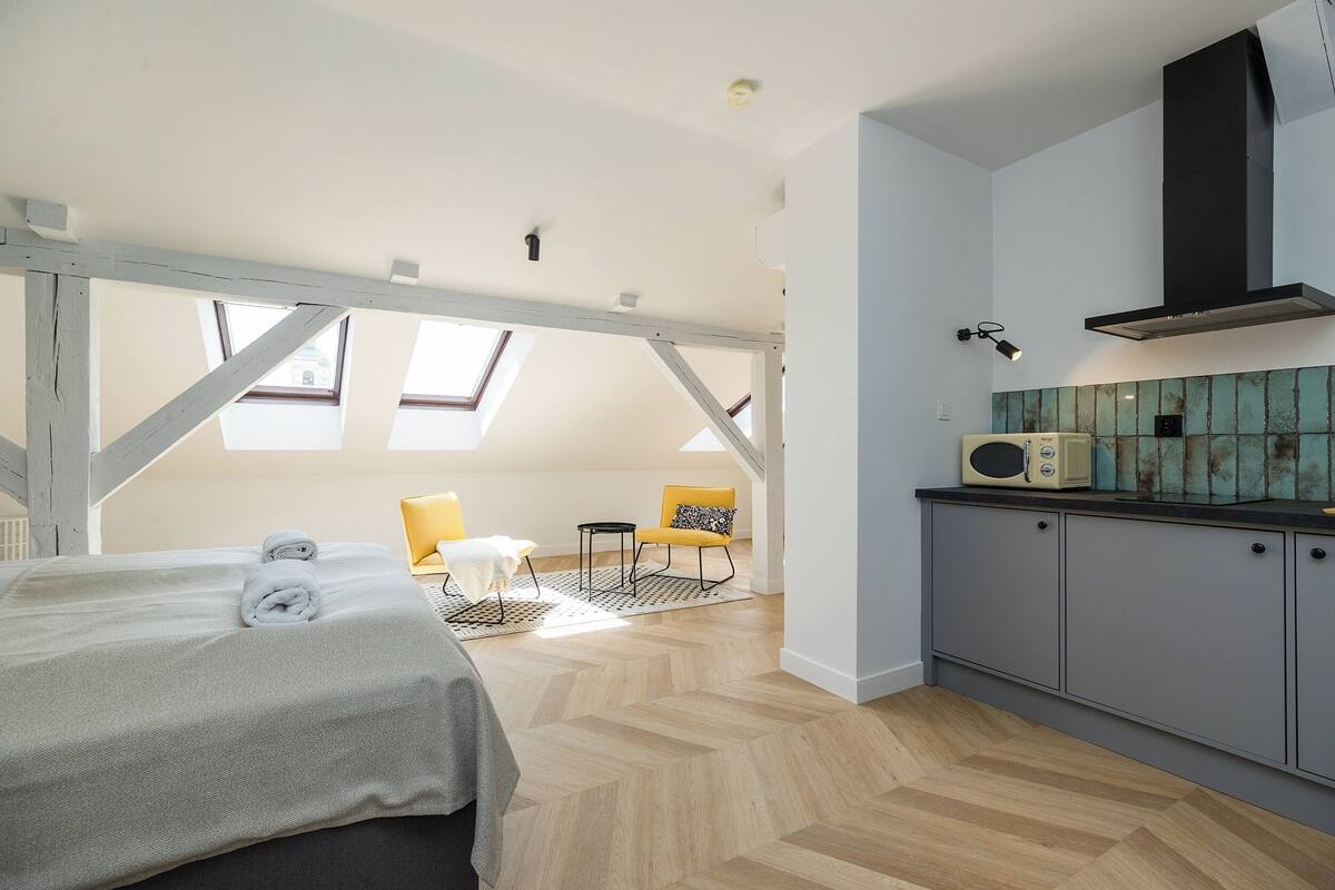A modern loft bedroom with a large bed, a small table, and a chair. There is a kitchenette with a microwave and a stove hood. The room has a sloped ceiling with skylights and a herringbone-patterned floor.