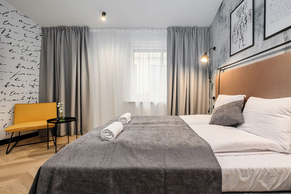 A hotel room with a bed, a chair, a small table, and a wall with a script-like pattern.