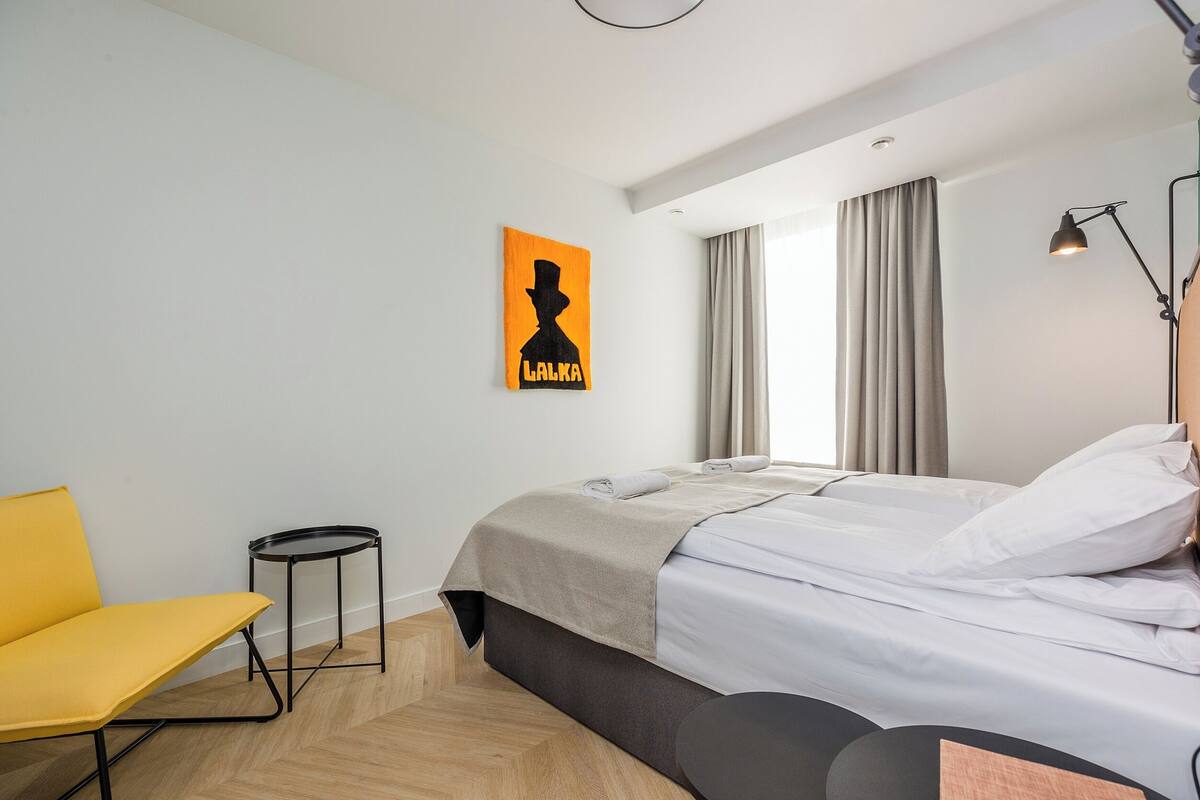 A modern hotel room with a bed, a small round table, a yellow chair, and a painting on the wall.