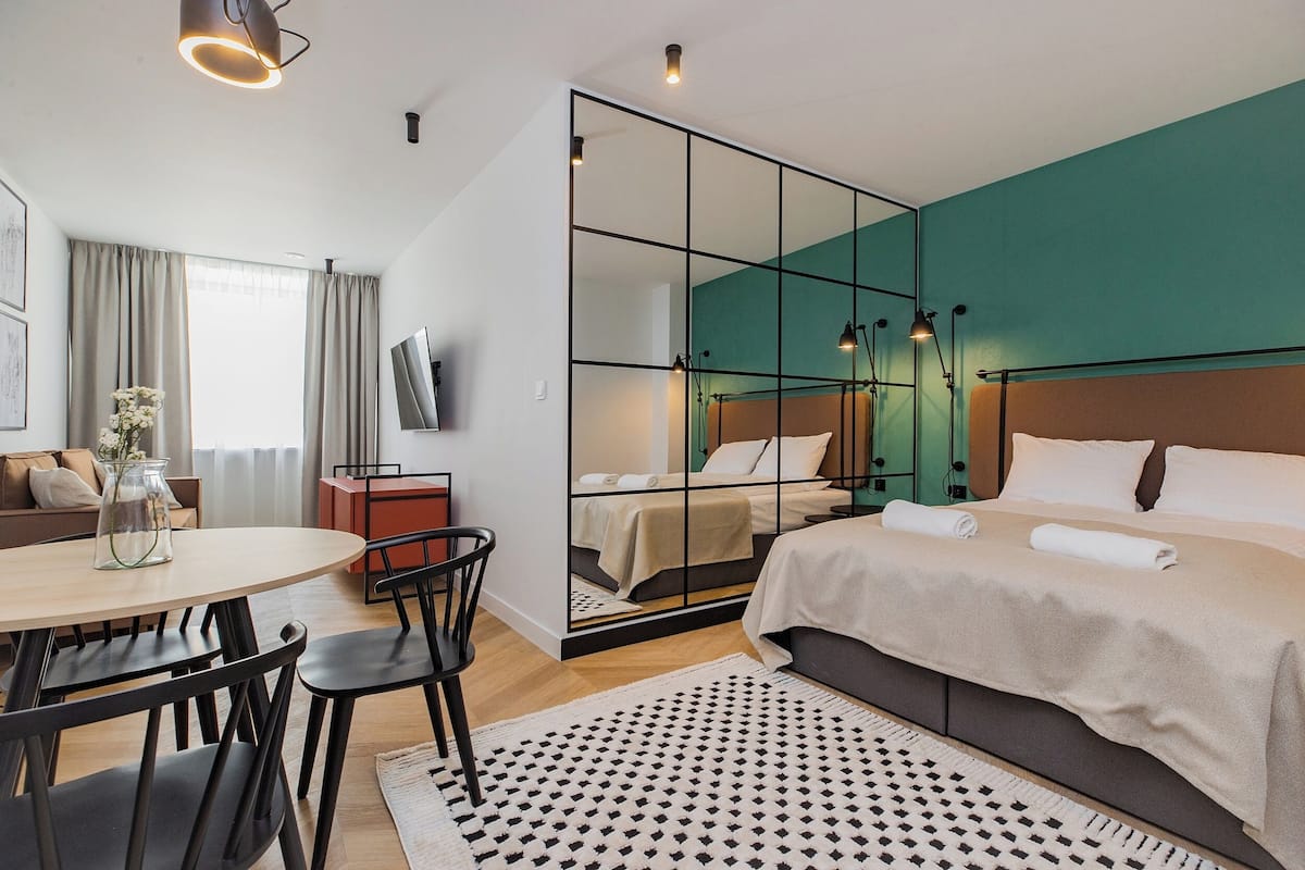 A modern hotel room with a large bed, a dining area with a table and chairs, a TV, and a large mirror.