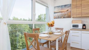 A dining area with a table set for four, wooden chairs, a vase with flowers, and a view of greenery through large windows. - Bright Apartment with a Balcony | Lake View, Bicycles (Katowice)