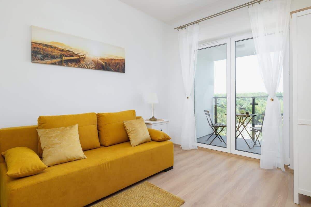 A bright room with a yellow sofa, a painting on the wall, and a balcony with a table and chairs.