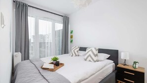 Een moderne slaapkamer met een groot bed, grijze gordijnen, een nachtkastje met een lamp en een raam met uitzicht op de stad.