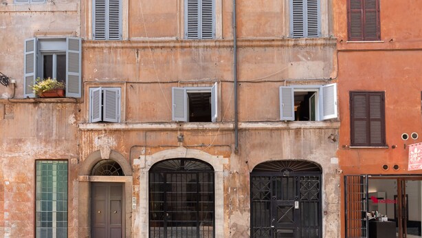 Un bâtiment doté de nombreuses fenêtres, dont certaines sont équipées de volets, d’une grande porte en arc de cercle et d’une petite fenêtre ornée d’une plante.