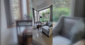 A room with a stone wall, a window with a view of greenery, a sofa, and a small table with a lamp.