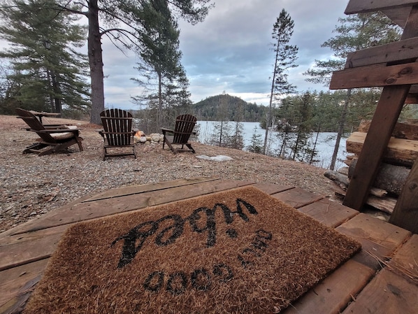Um terraço com cadeiras Adirondack, uma lareira e vista para um lago rodeado por árvores.