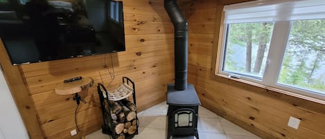 A cozy room with a wooden interior, a television mounted on the wall, a wood-burning stove, and a window with a view of trees.