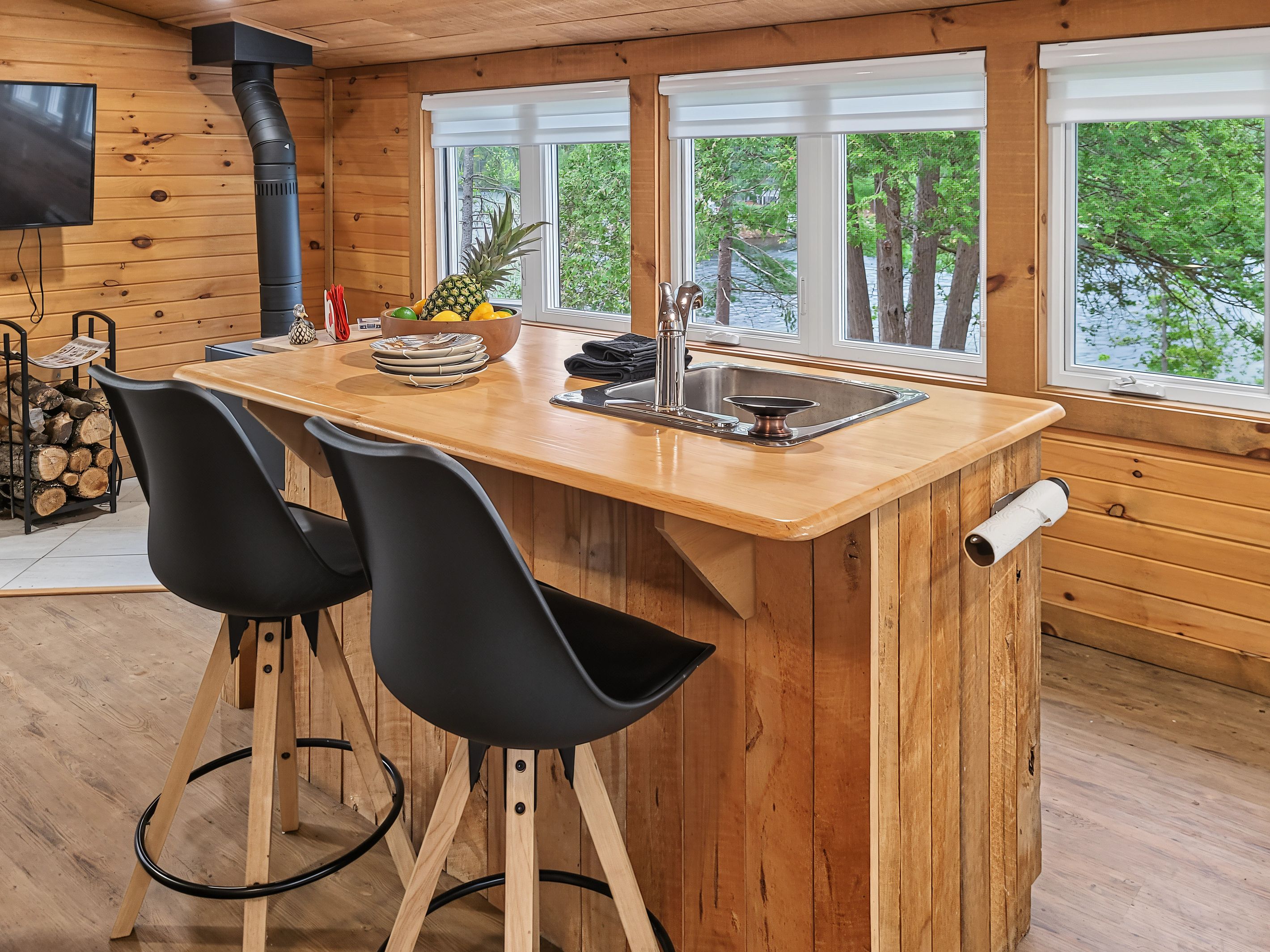 A wooden cabin interior with a kitchen island, two chairs, a sink, and a window overlooking greenery.