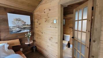A wooden cabin interior with a bed, a painting, a vase with flowers, a toilet, and a door with frosted glass panels.