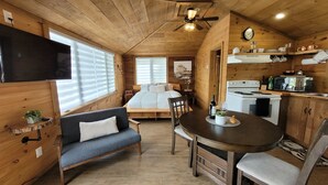 A cozy cabin-style room with a wooden interior, a dining table with chairs, a sofa, a bed, and a kitchenette.