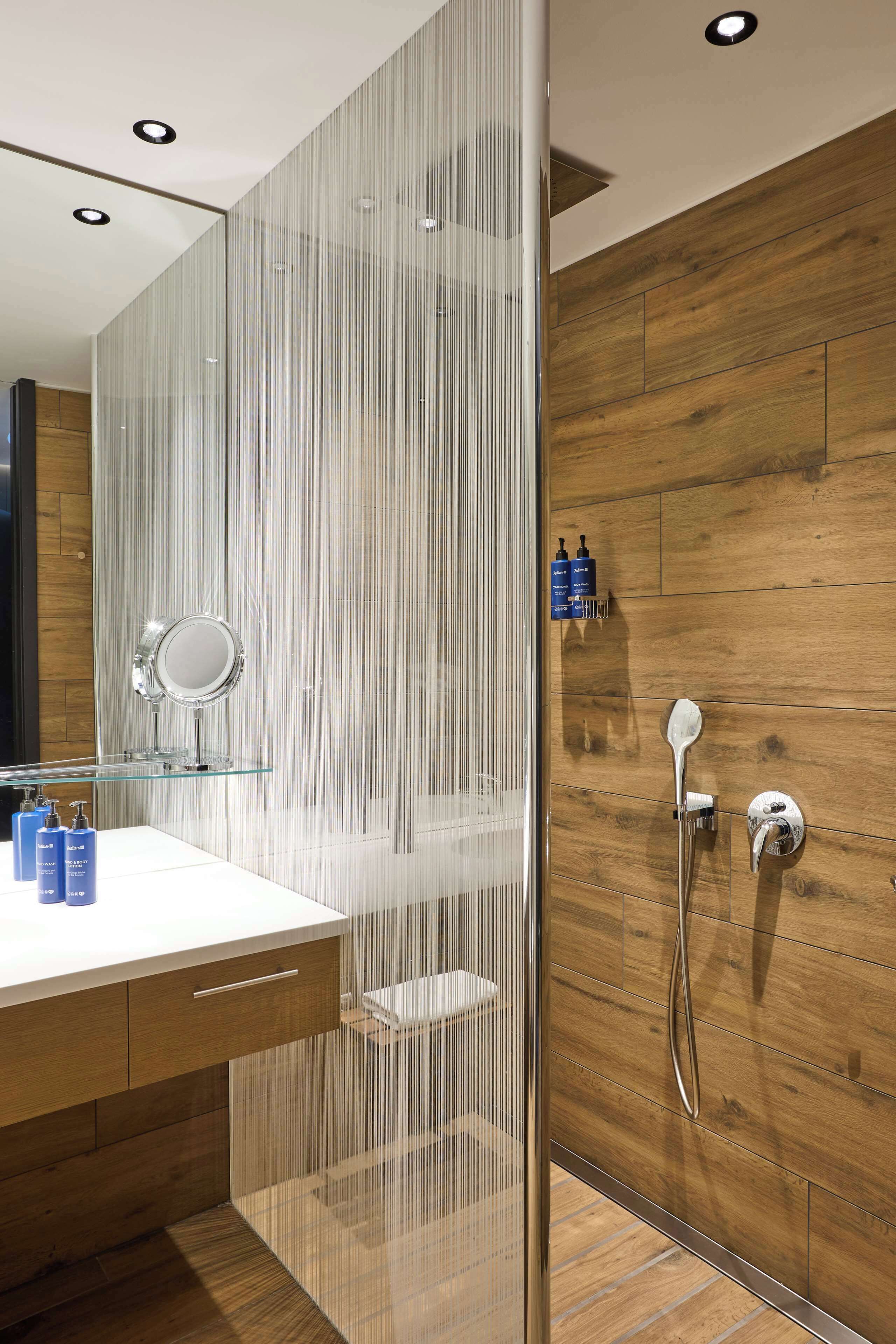 A modern bathroom with a glass shower enclosure, wooden wall paneling, a white vanity with a mirror, and toiletries on the counter.