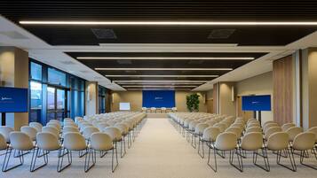 Un salón de conferencias con filas de sillas mirando hacia un escenario, ventanas amplias y un cartel azul con texto.