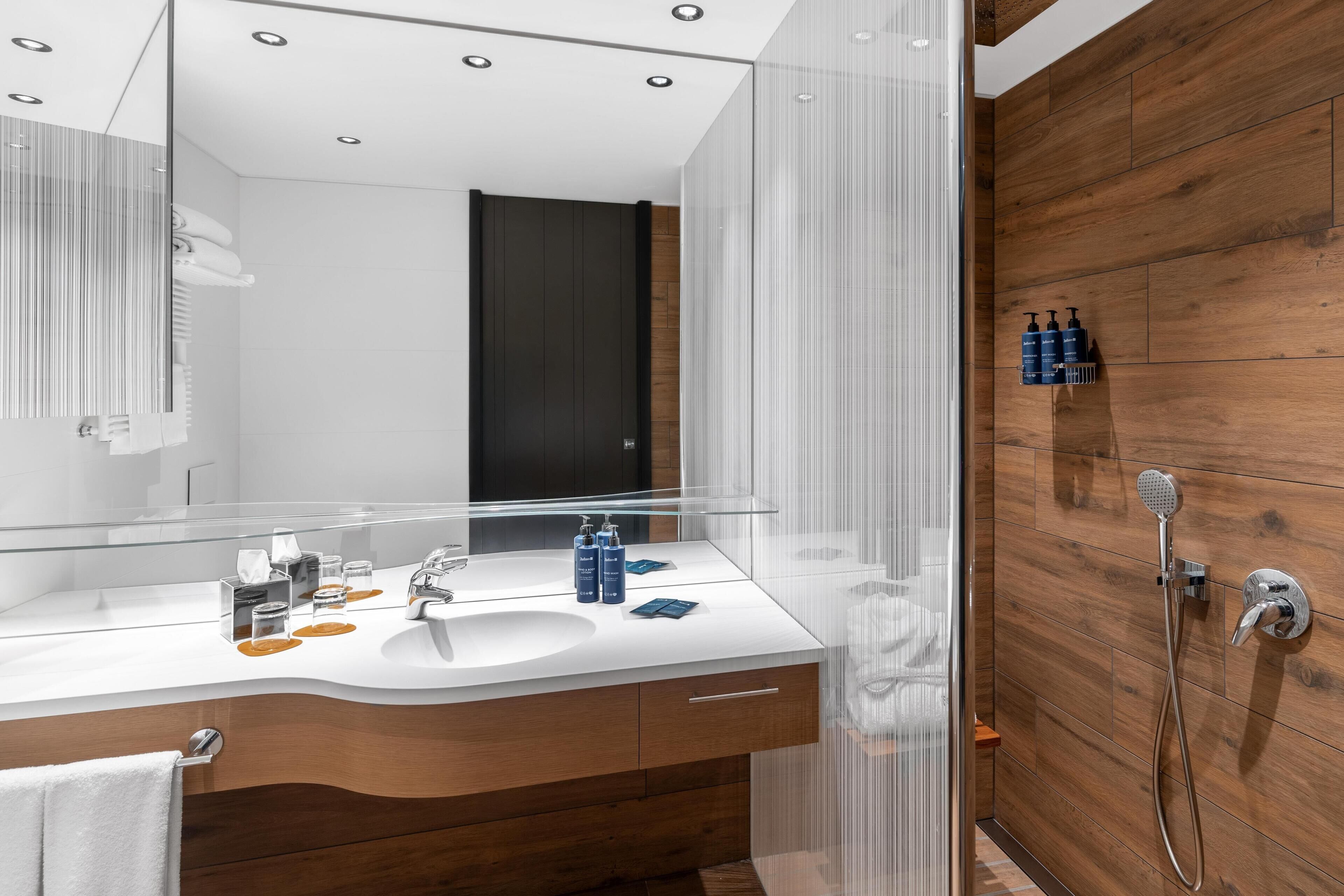 A modern bathroom with a wooden wall, a glass shower enclosure, and a white sink with toiletries.