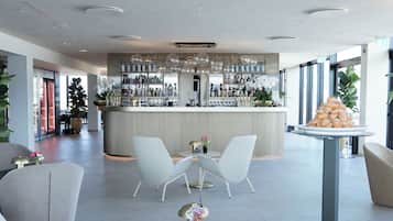 Un área de bar moderna con sillas blancas y una mesa redonda, una barra con varios botellas y una planta grande en maceta.