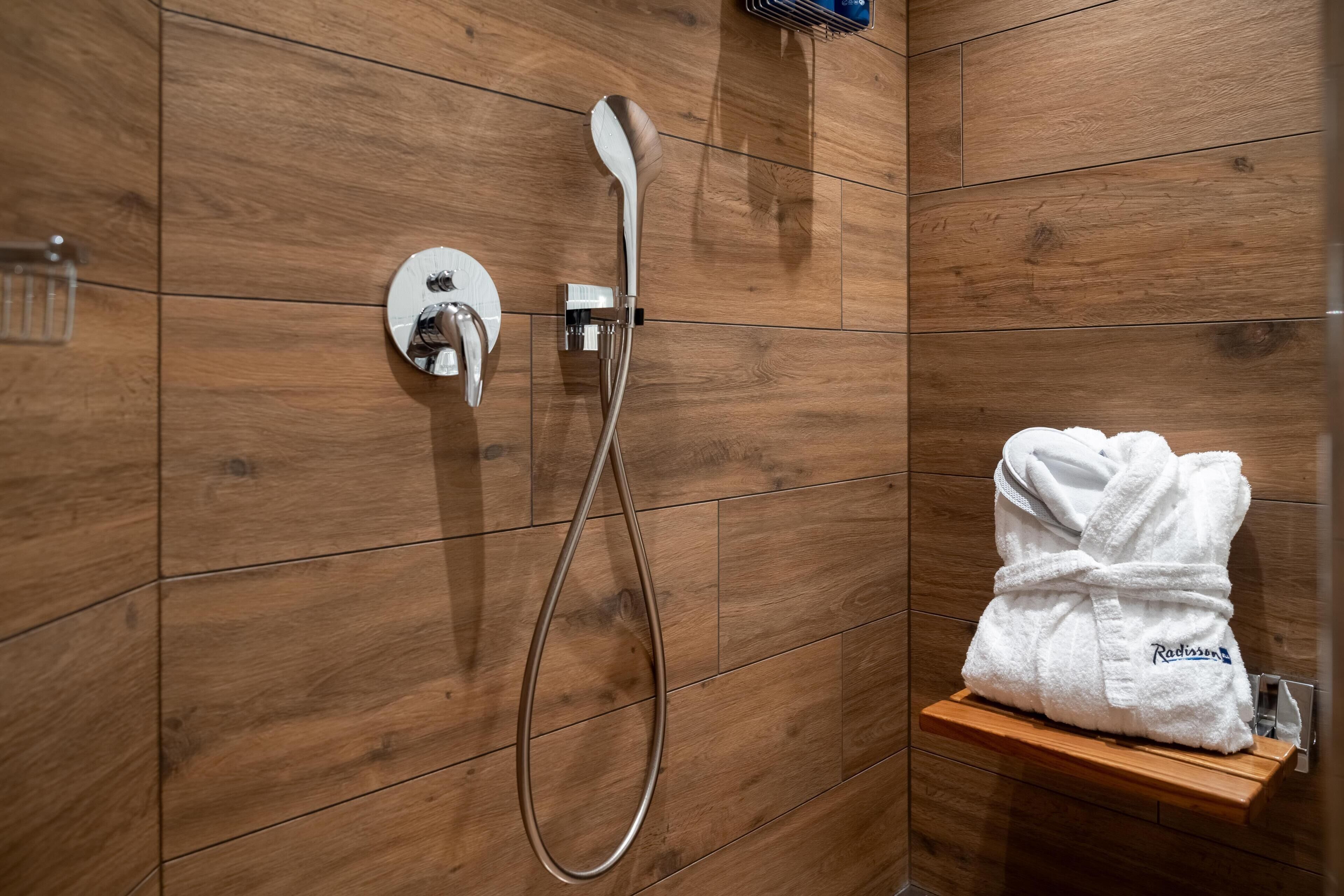 Ducha con cabezal de mano y panel de control en la pared. Un albornoz blanco está colocado en un estante de madera.