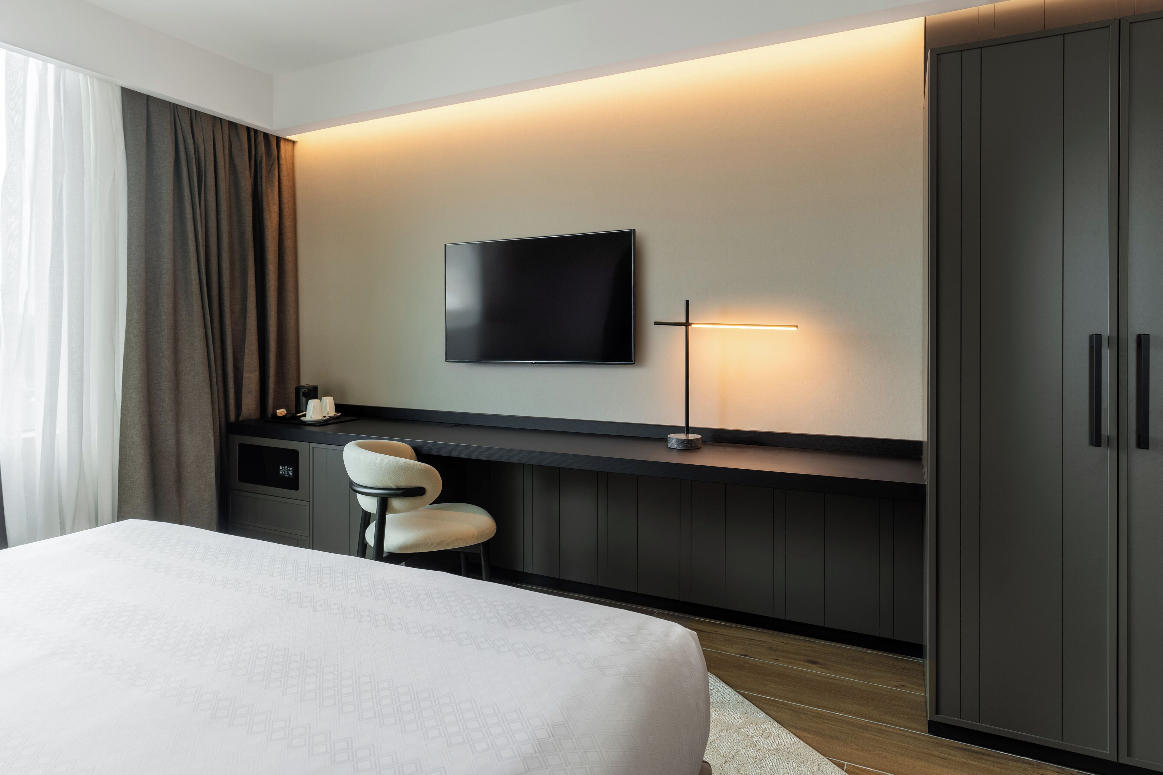 A modern hotel room with a large bed, a desk with a chair, a flat-screen TV mounted on the wall, and a wardrobe.