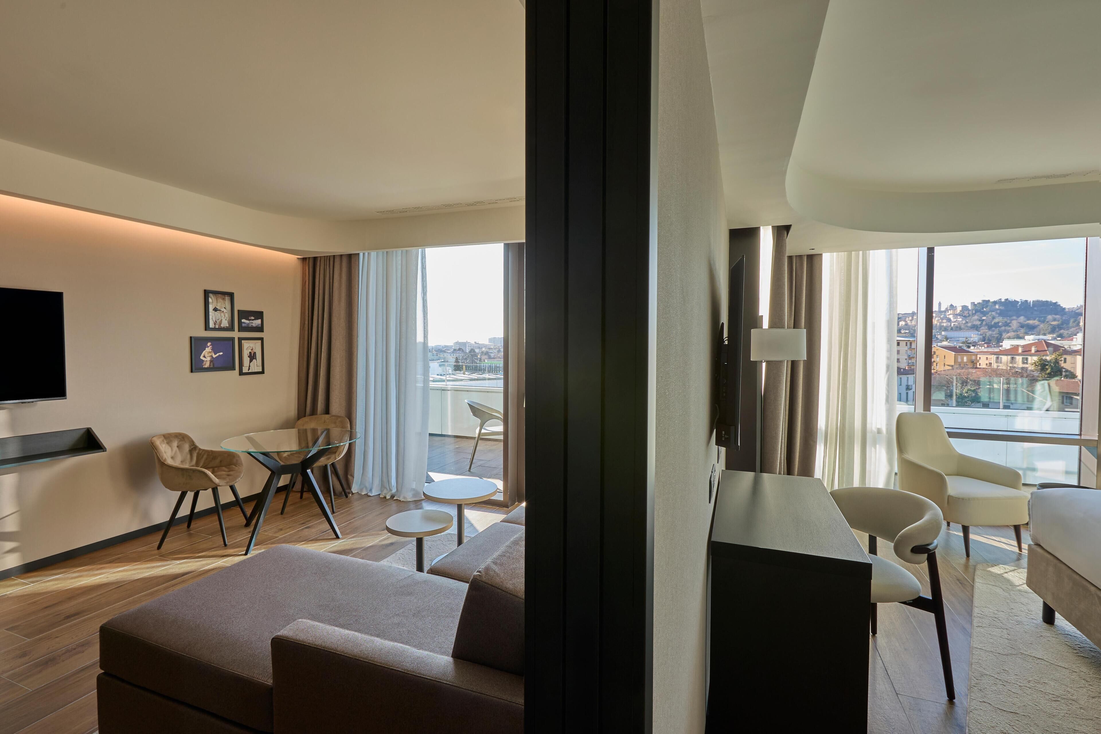 A modern hotel room with a large window offering a city view, a flat-screen TV mounted on the wall, a sofa, a dining table with chairs, and a small round table.