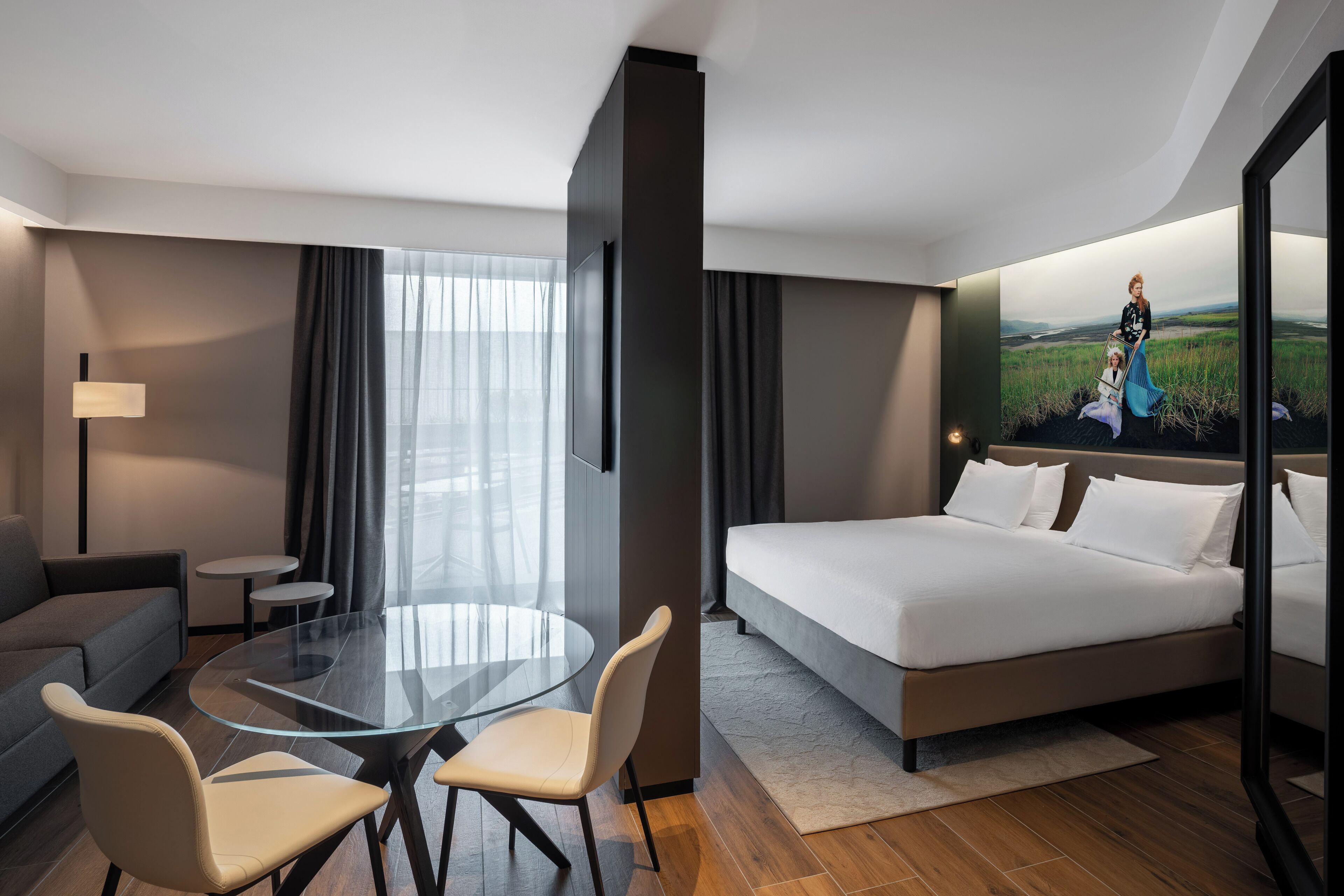 A modern hotel room with a glass dining table, a sofa, a bed, and a large window with a view of a field.