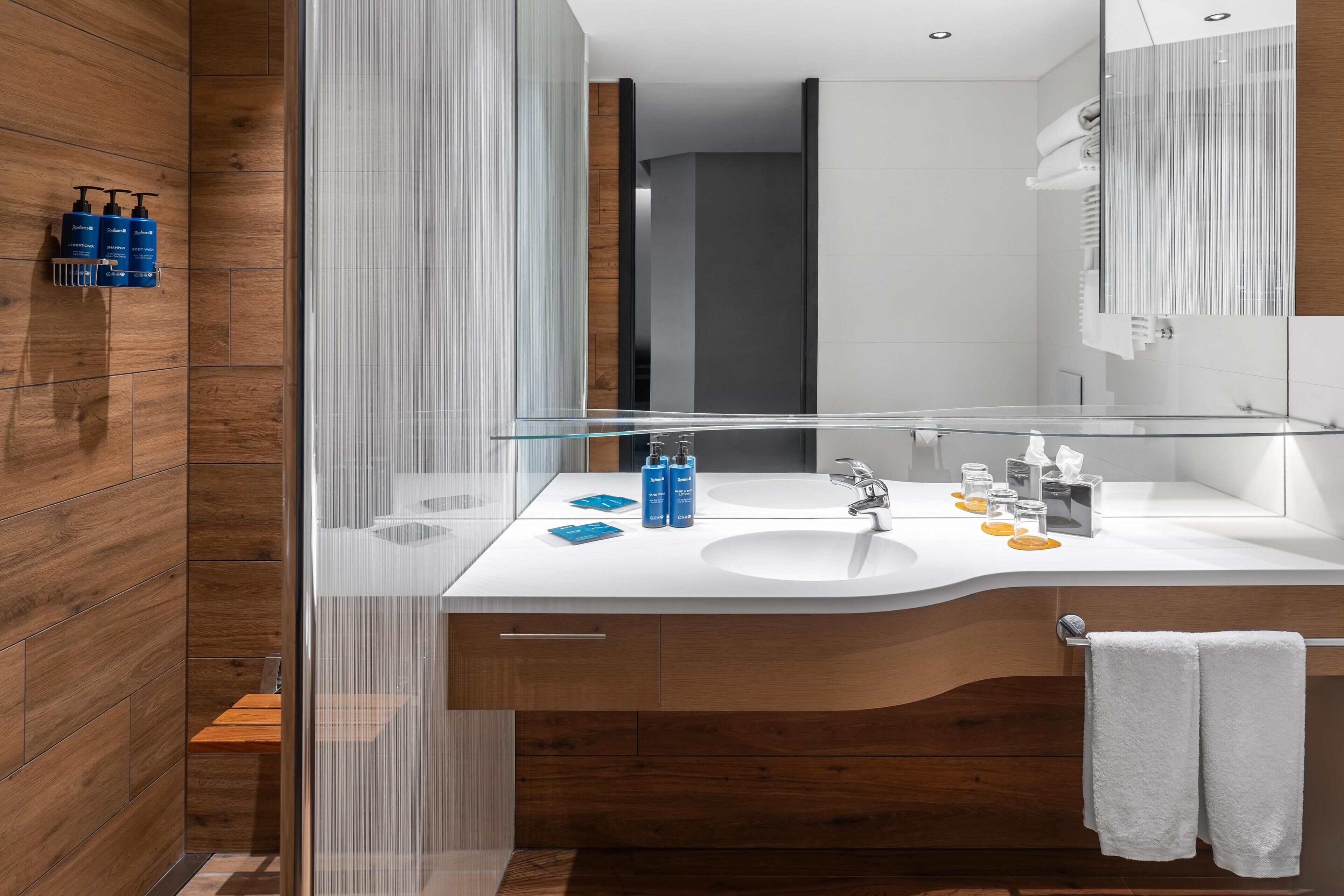 A modern bathroom with a wooden wall, a glass shower enclosure, and a white sink with toiletries.