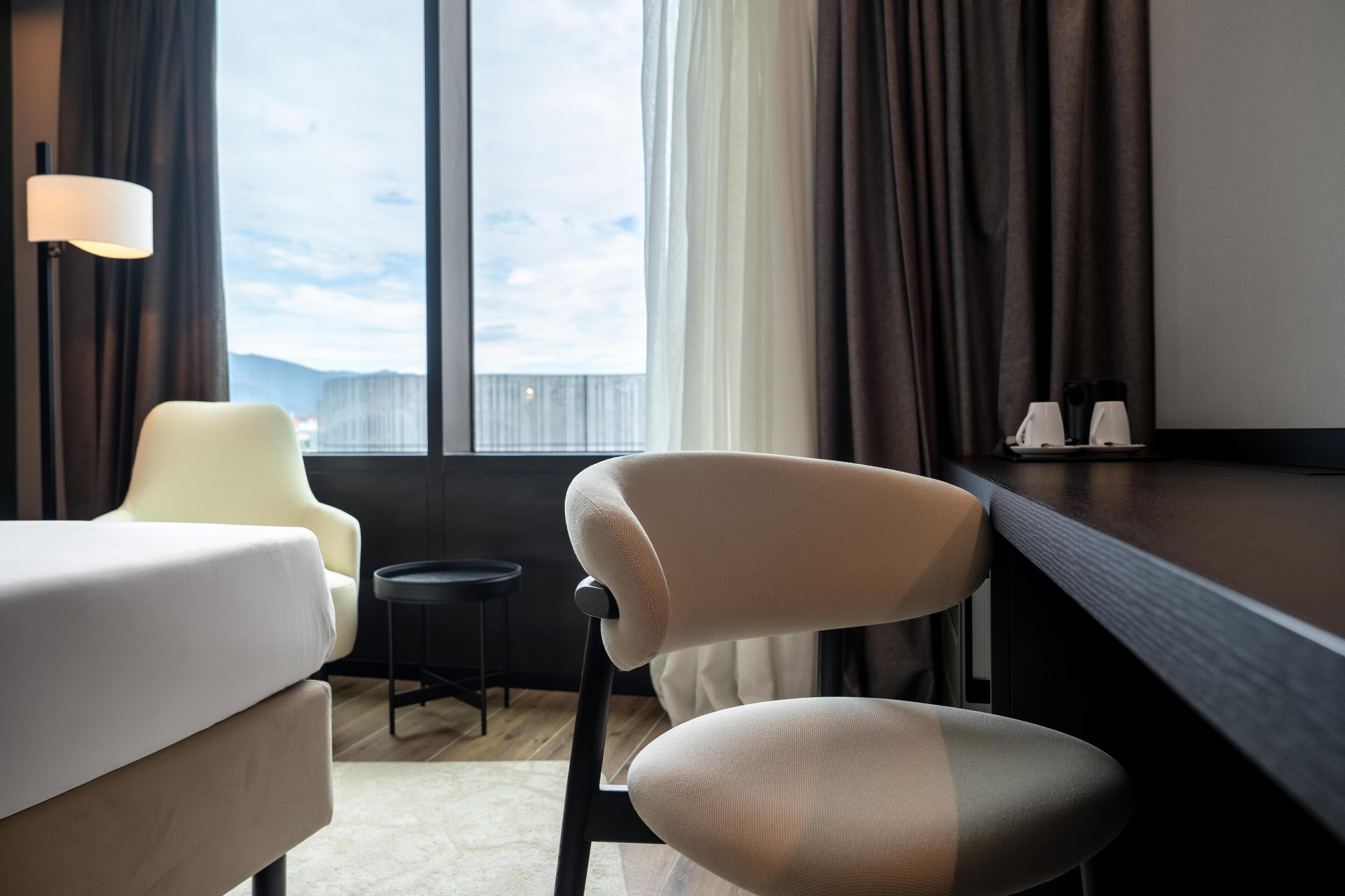 A hotel room with a bed, a chair, a small table, a lamp, and a view of the outdoors.