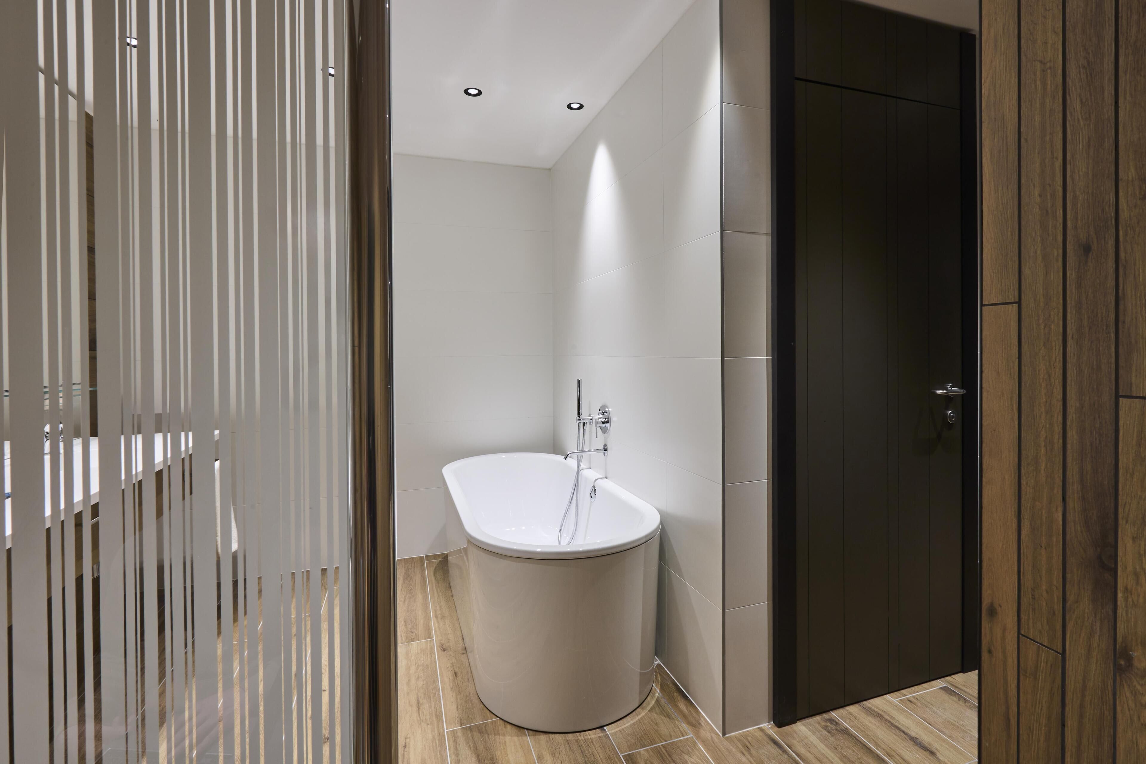 A modern bathroom with a freestanding bathtub, wooden flooring, and a dark wooden door.