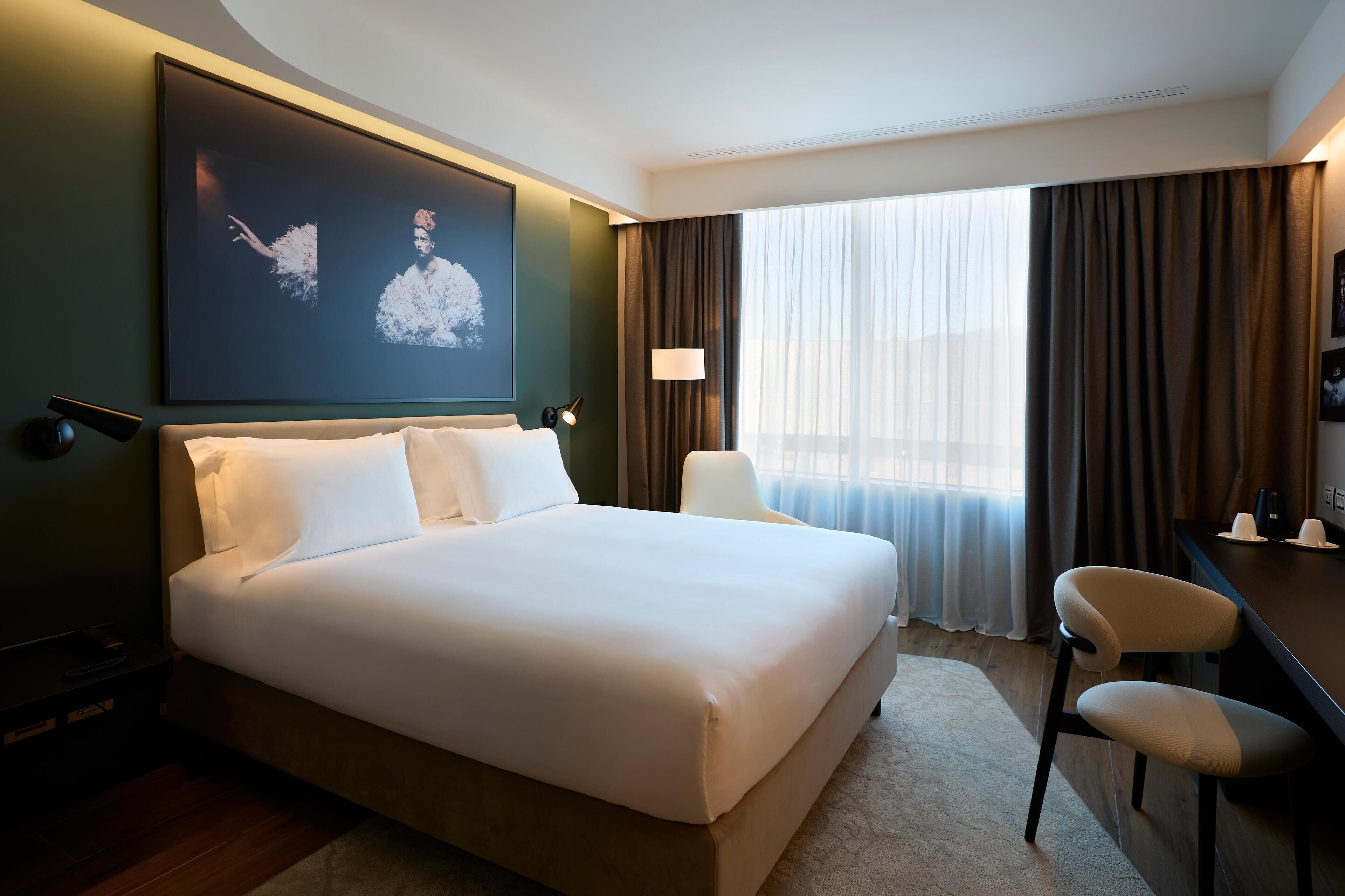 Habitación de hotel con una cama grande, un escritorio, una silla, un televisor y una ventana con cortinas.