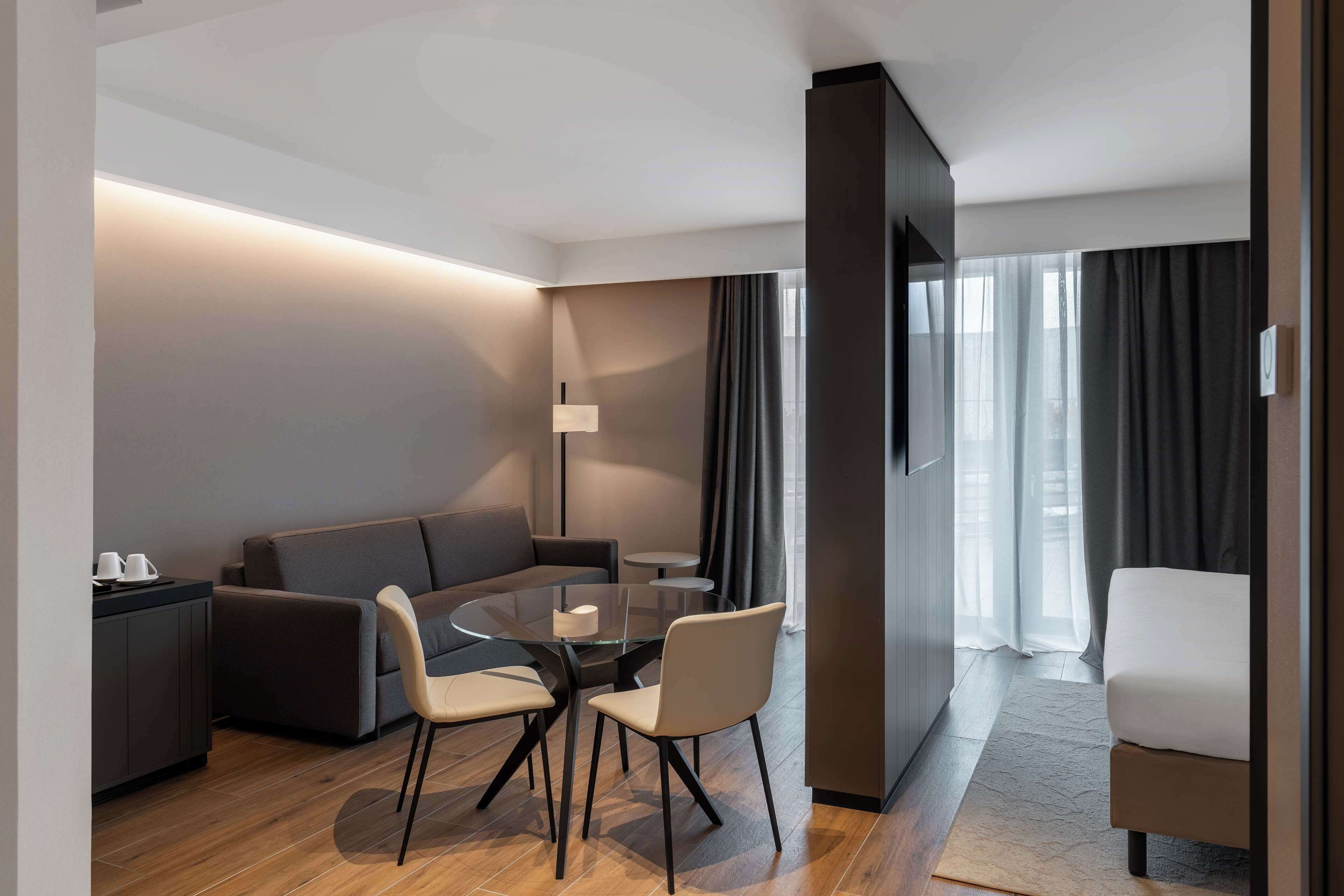 A modern hotel room with a round dining table, two chairs, a sofa, a bed, and a TV.