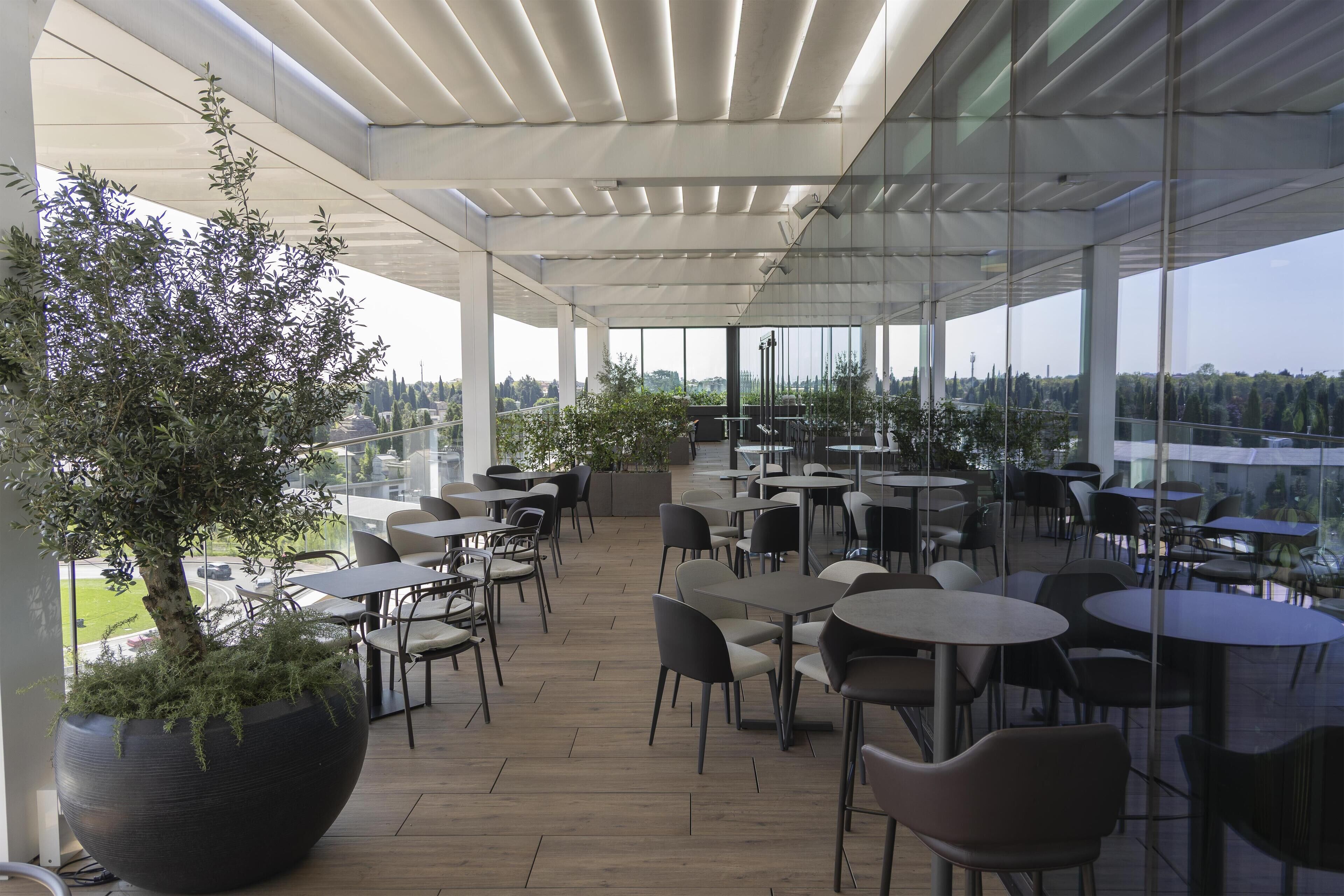 An outdoor café area with round tables and chairs, large potted plants, and a view of the cityscape through large windows.