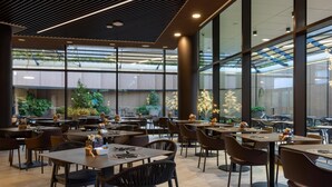 Een modern restaurant met grote ramen, kamerplanten en diverse zithoeken.