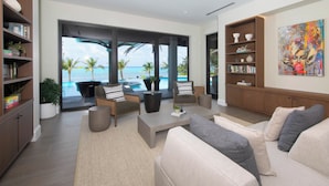 A spacious living room with a large window overlooking a pool and ocean, furnished with a sofa, chairs, and a coffee table.