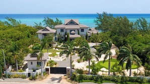 A large, multi-story house with a well-maintained lawn and palm trees, located near a beach with clear blue waters.