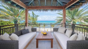 A covered outdoor seating area with a wooden table and two sofas, overlooking a beach with palm trees and the ocean.
