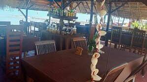 Un ristorante sulla spiaggia con tavoli e sedie di legno, un tetto di paglia e bottiglie sul bancone.
