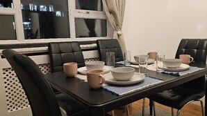 Een eettafel is gedekt met borden, kommen en bekers, omringd door zwarte stoelen, in een kamer met een raam en uitzicht op gebouwen.