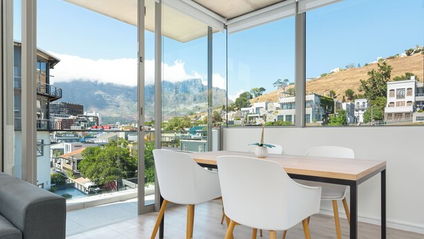 A modern dining area with a large window offering a view of a cityscape and mountains. - Luxury 2 Bedroom Mountain Views (Cape Town)