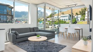 A modern living room with a grey sectional sofa, a round coffee table with a plant, a black and white patterned rug, and a dining area with white chairs and a wooden table. - Luxury 2 Bedroom Mountain Views (Cape Town)