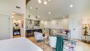 Una sala de estar moderna y bien iluminada con un sofá, sillas, una mesa de centro y una cocina al fondo.