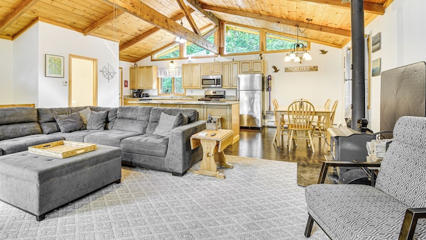 A spacious living area with a gray sectional sofa, a wooden coffee table, and a patterned armchair. A kitchen with modern appliances and a dining area with wooden chairs is visible in the background. - Peaceful Highland Lake Home w/ Kayaks, Dock Access (Bridgton)