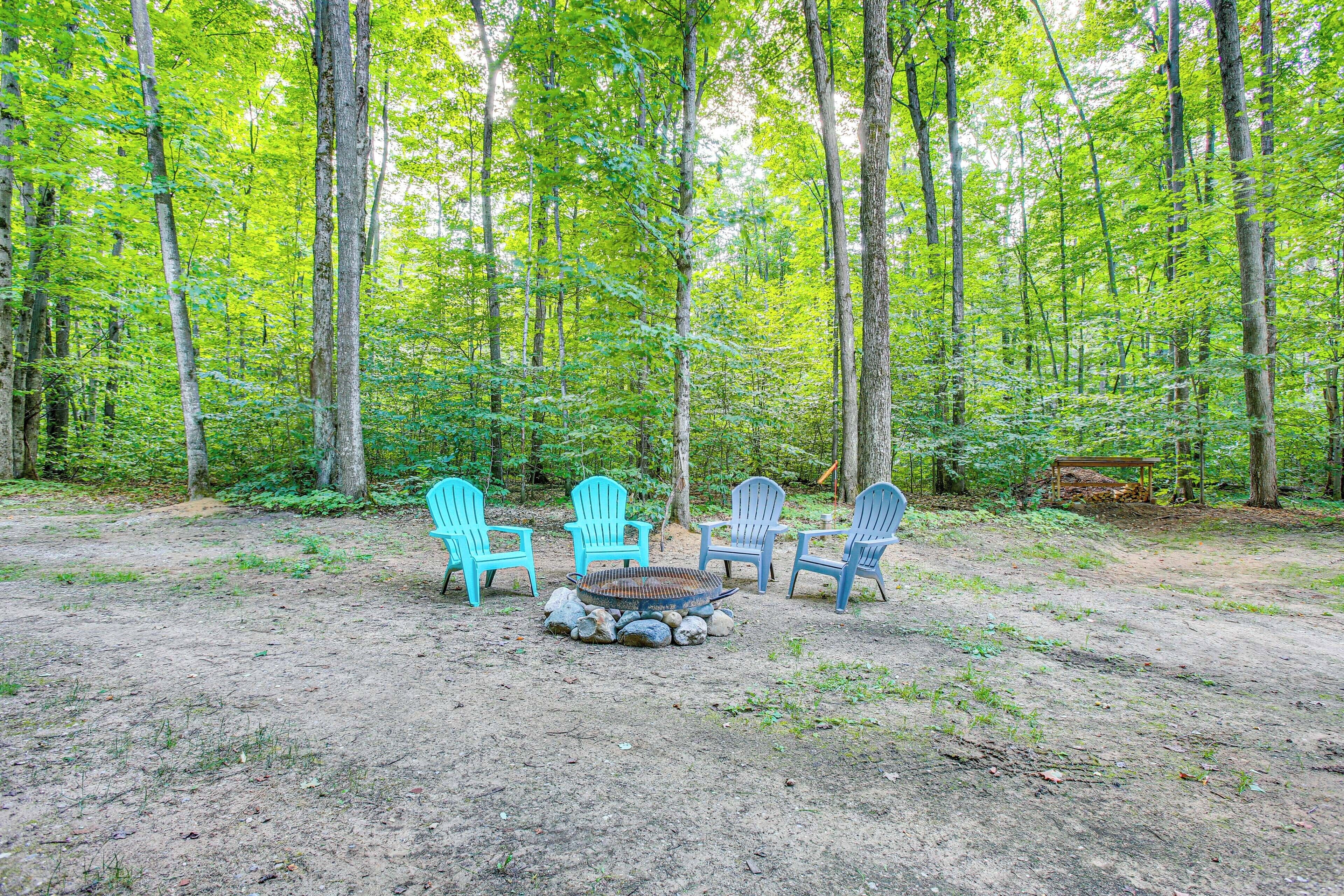 A forest clearing with a fire pit and four Adirondack chairs.