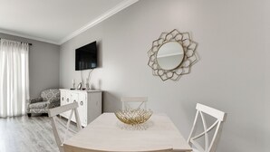 A dining area with a wooden table, white chairs, a decorative bowl, a television, and a patterned armchair.